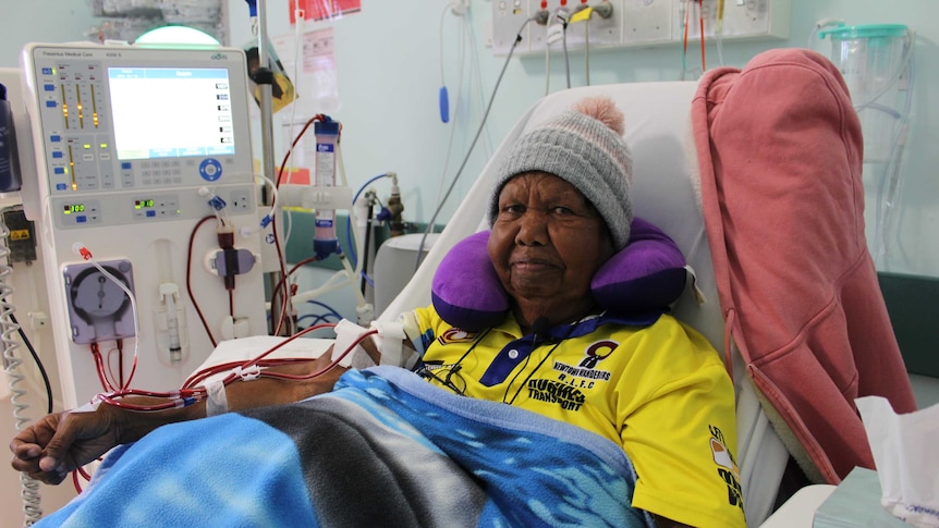 A woman, wearing a beanie and a blanket, receives dialysis treatment in a hospital bed