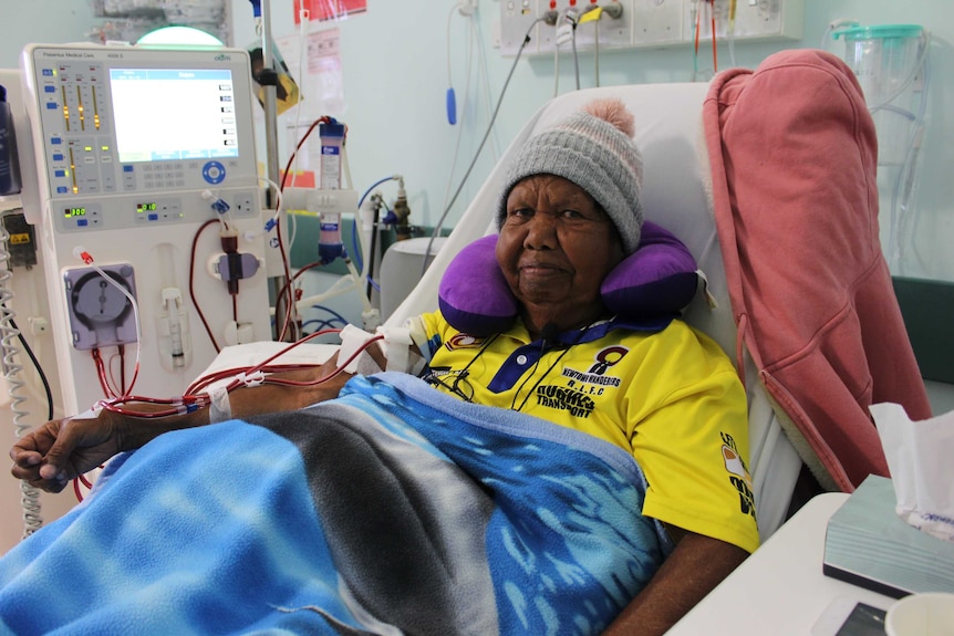 A woman, wearing a beanie and a blanket, receives dialysis treatment in a hospital bed