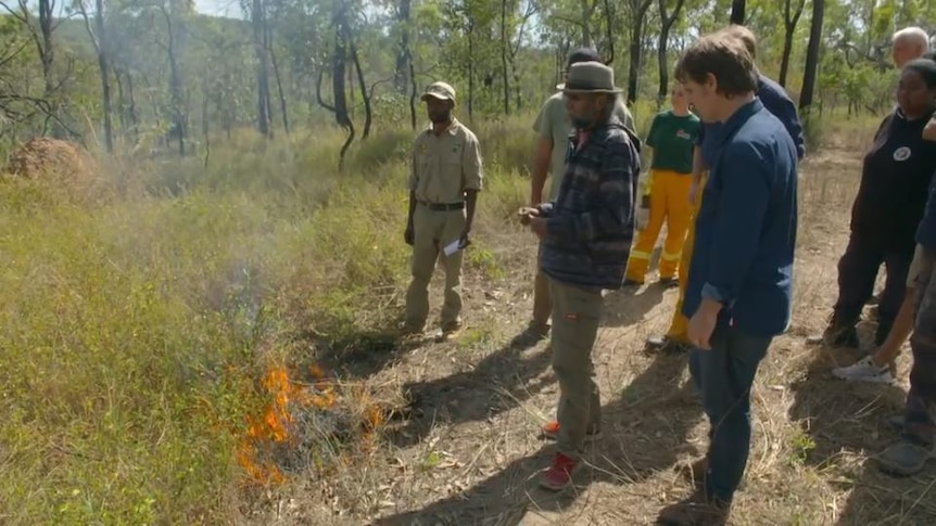 Craig Reucassel stands with Indigenous men beside small fire in grassland