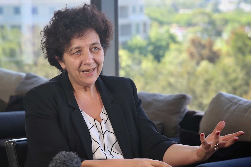 Madame Vidal is sitting on a chair in a hotel, gesturing with her hands. Trees can be seen out of the window.