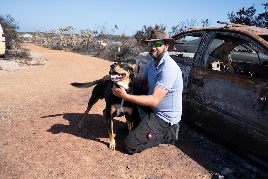 A man squats beside his dog and his burnt-out car