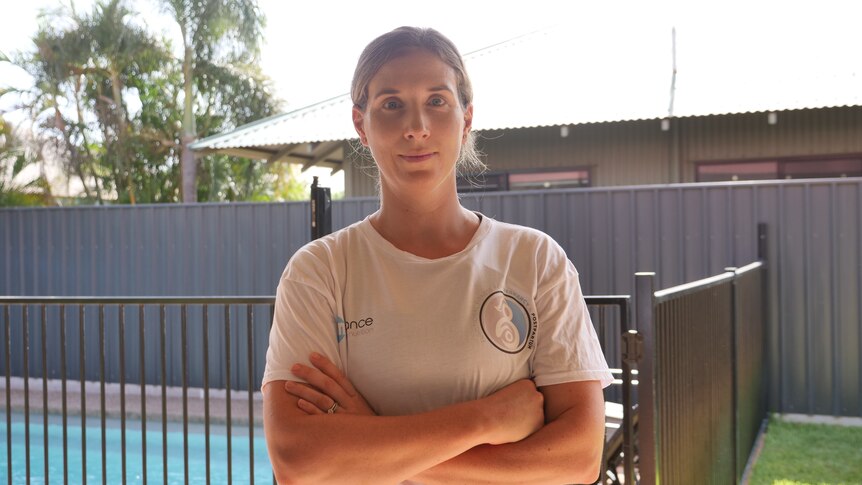 A woman in a white shirt stands in front of a fenced pool