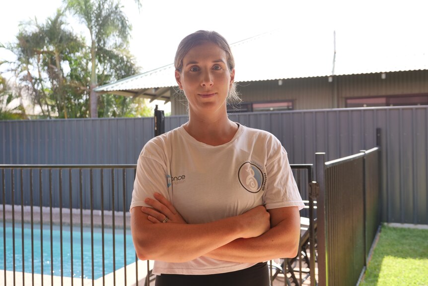 A woman in a white shirt stands in front of a fenced pool