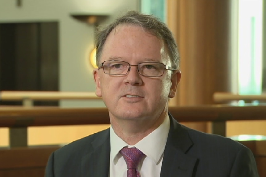 Middle aged bespectacled white man wearing a suit and tie sitting in an office foyer