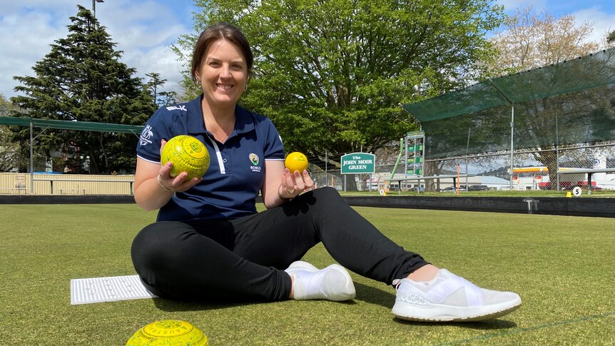 A woman sits on a bowling green, smiling as she holds two lawn bowls.