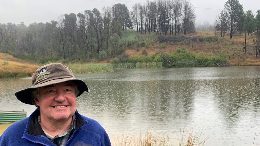 A man in a hat and blue jumper stands in front of a dam on a rainy day