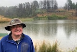 A man in a hat and blue jumper stands in front of a dam on a rainy day