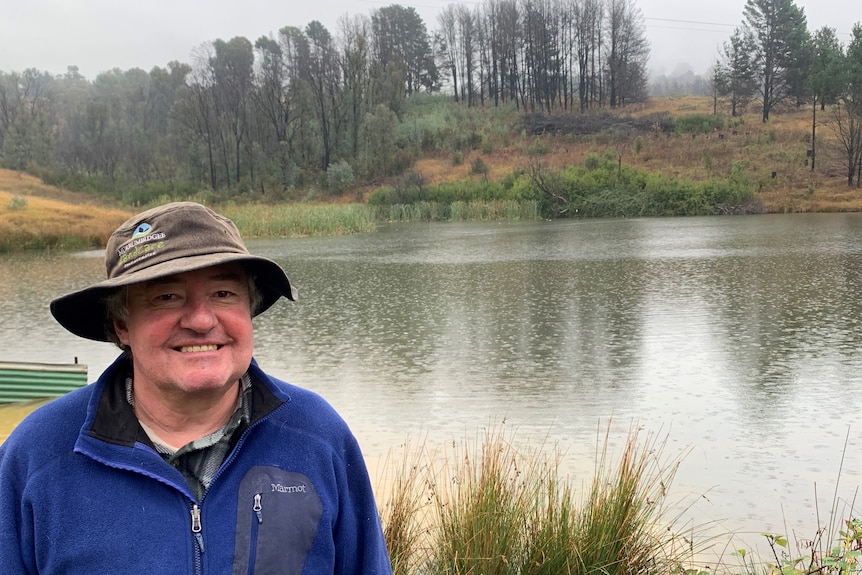 A man in a hat and blue jumper stands in front of a dam on a rainy day