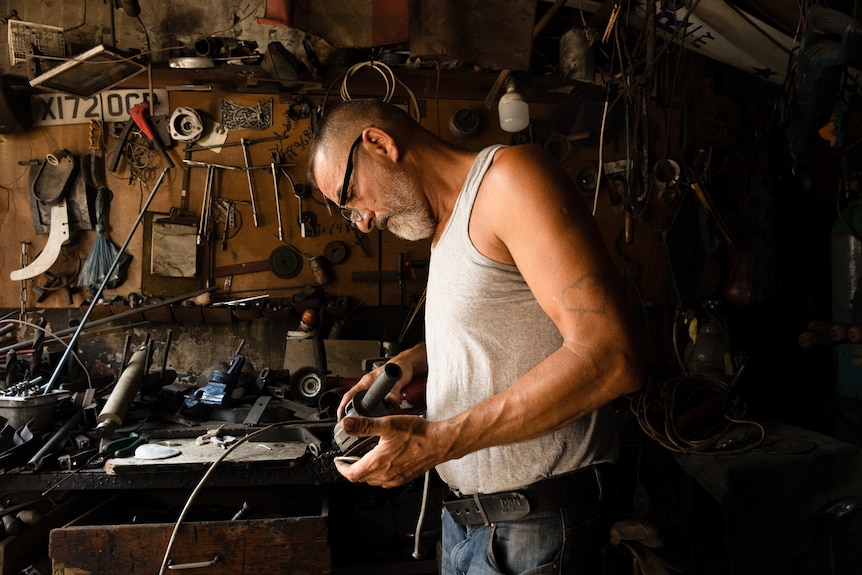 A man wears a white singlet as he leans over a tool in his hands in a shed.