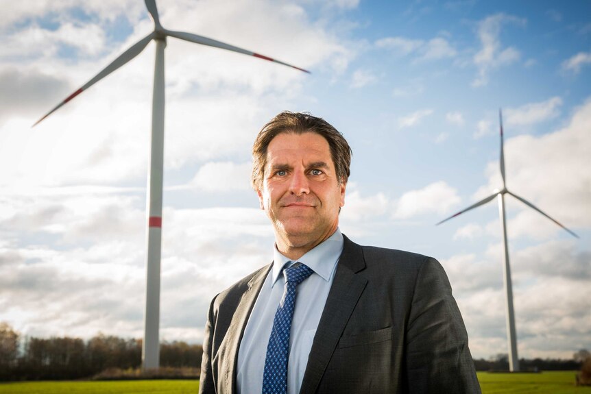 A man wearing a suit stands in front of two wind turbines on sunny day