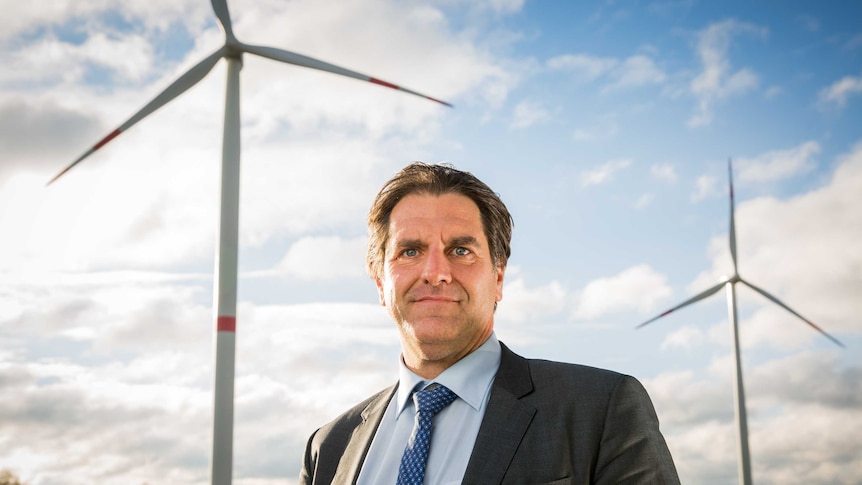 A man wearing a suit stands in front of two wind turbines on sunny day