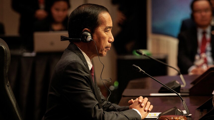 Joko Widodo, seen from side profile, sits at a desk with his hands folded and headphones on 