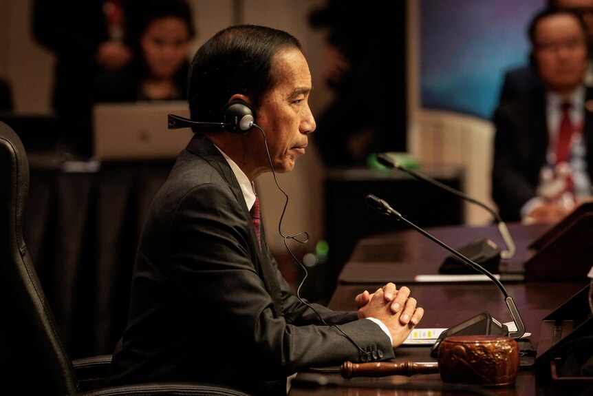 Joko Widodo, seen from side profile, sits at a desk with his hands folded and headphones on 