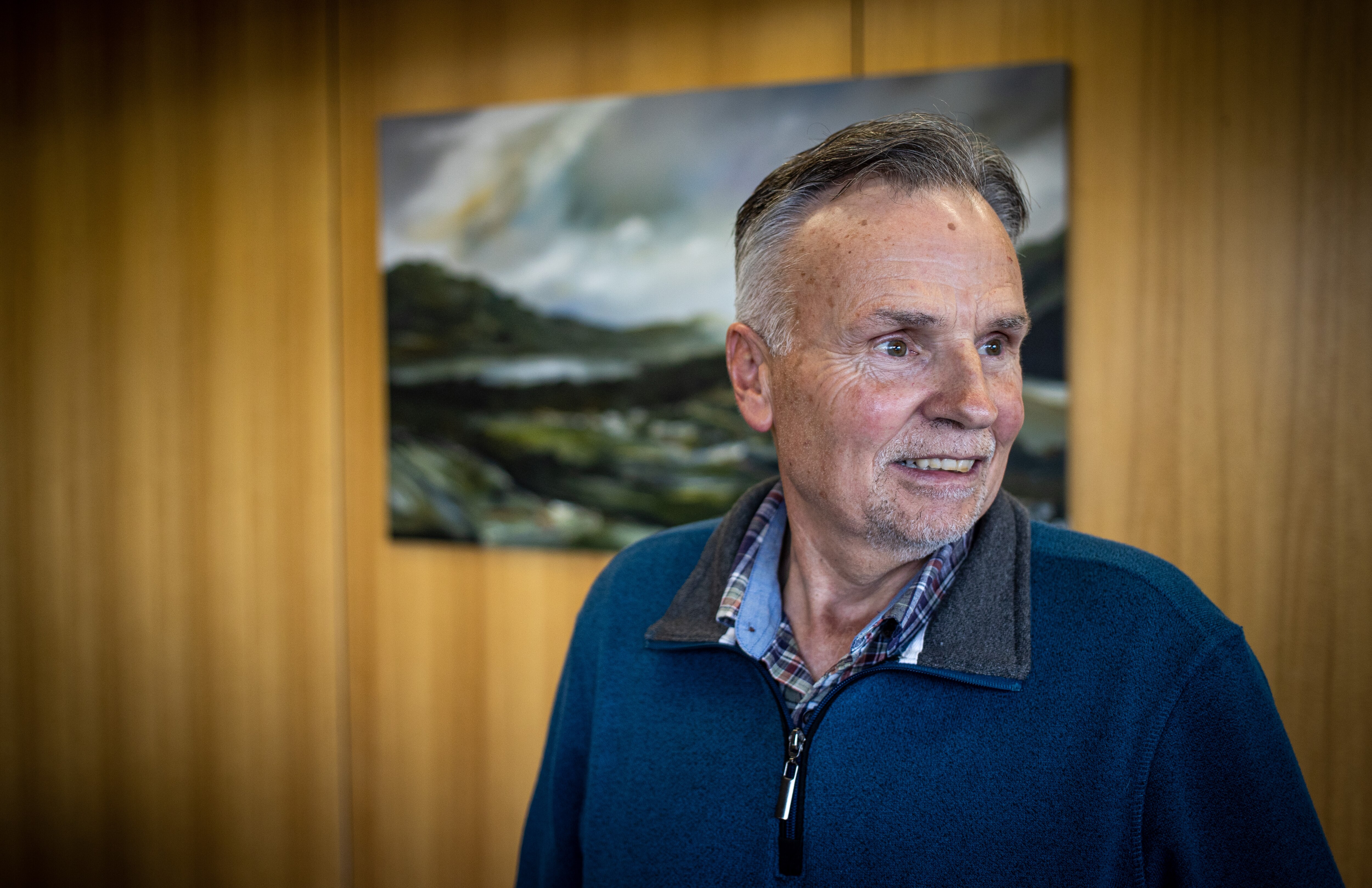 Jon Moore stands in front of a wooden wall with a landscape painting haning on it, he has his head turned to one side