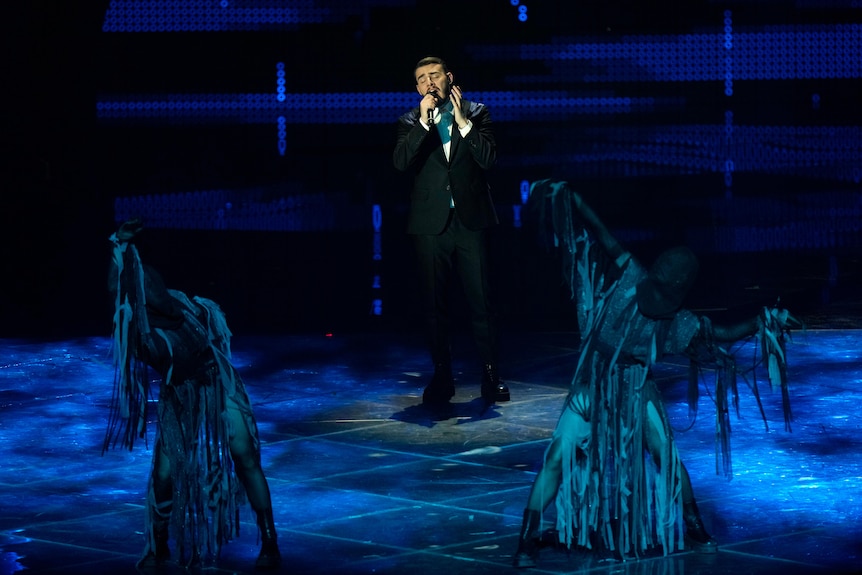 A man in a dark suit sings with his eyes closed on a blue-lit stage, as dancers move in front of him. 
