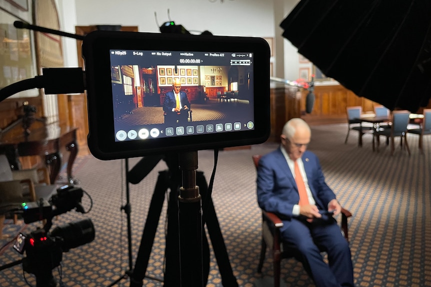 Malcolm Turnbull sits in a chair in a room. In the foreground, his image appears on a monitor screen.