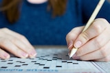 A hand holds a pencil, which hovers over the small black and white squares of an unfilled crossword puzzle.