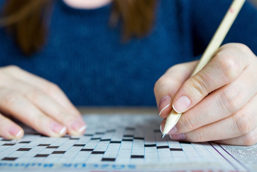 A hand holds a pencil, which hovers over the small black and white squares of an unfilled crossword puzzle.