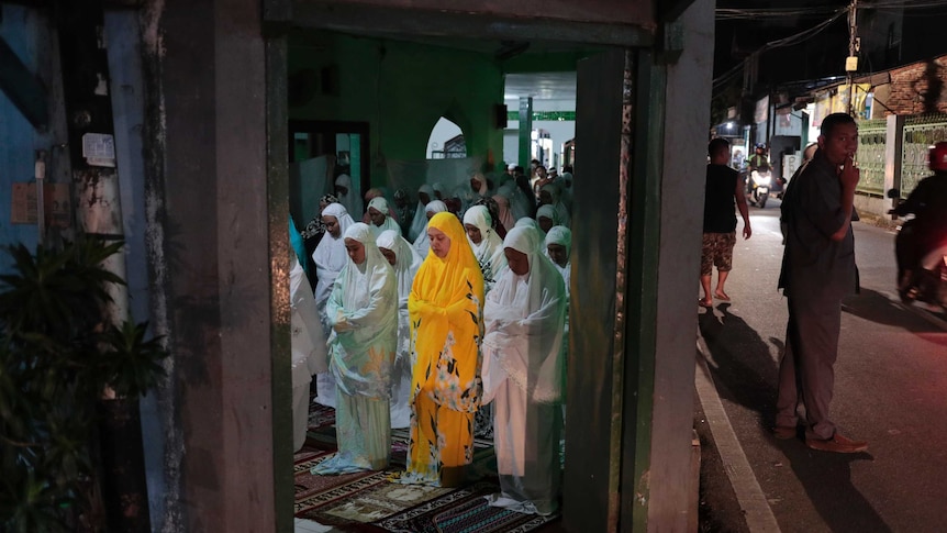 Women, mostly dressed in white, undertake evening prayers for Ramadan, while a man stands smoking.