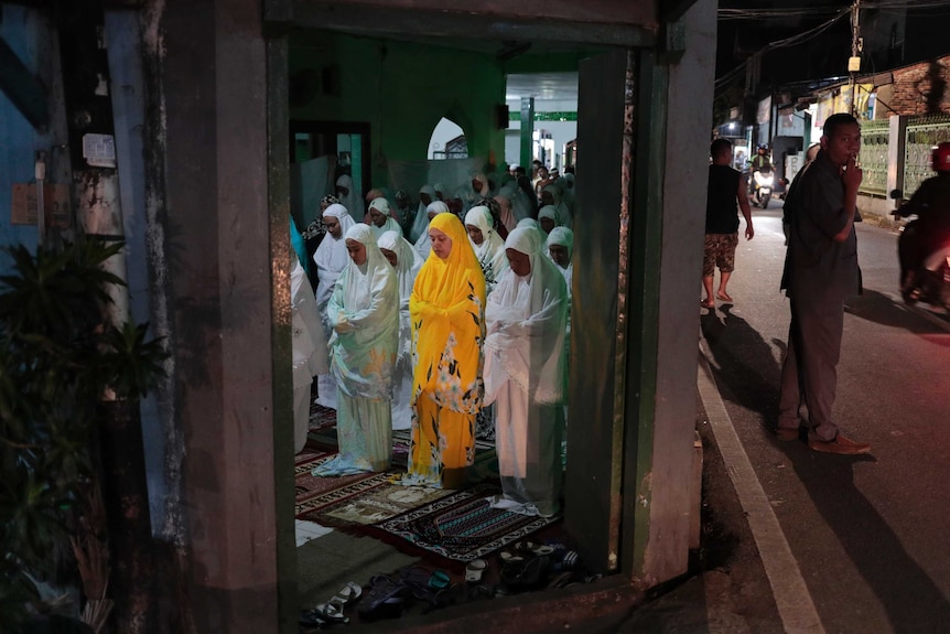 Women, mostly dressed in white, undertake evening prayers for Ramadan, while a man stands smoking.