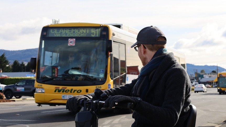 A man on a mobility scooter beside a suburban road, turning his body to look at a passing bus