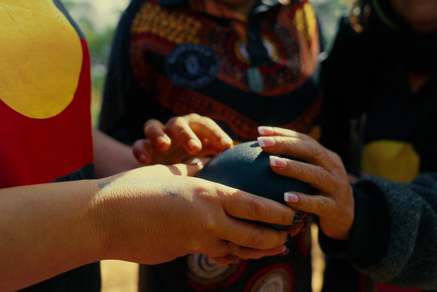 Three hands touch and hold a deep purple-coloured emu egg. They have a deep connection.