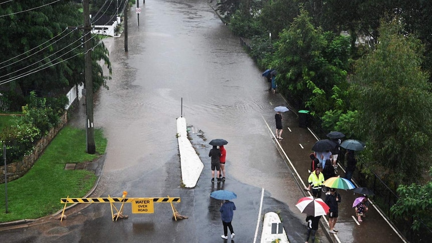 Flood waters spread across a road as a group of residents with umbrellas watch on.