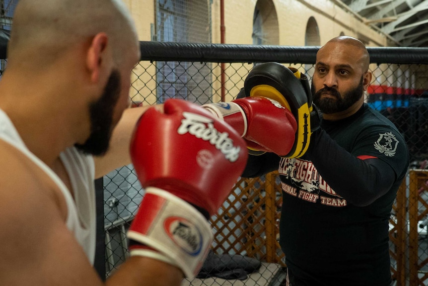 A man in singlet and wearing boxing gloves punches pads held by another man inside an MMA ring.