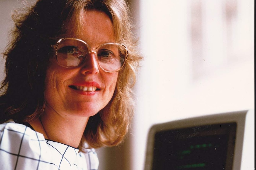 Helen Vorrath looking to camera in front of a 1980s personal computer with green text on the screen