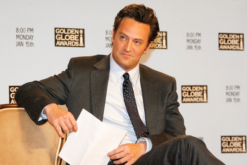 Matthew Perry sitting down wearing a suit and tie sits on a chair holding a piece of paper