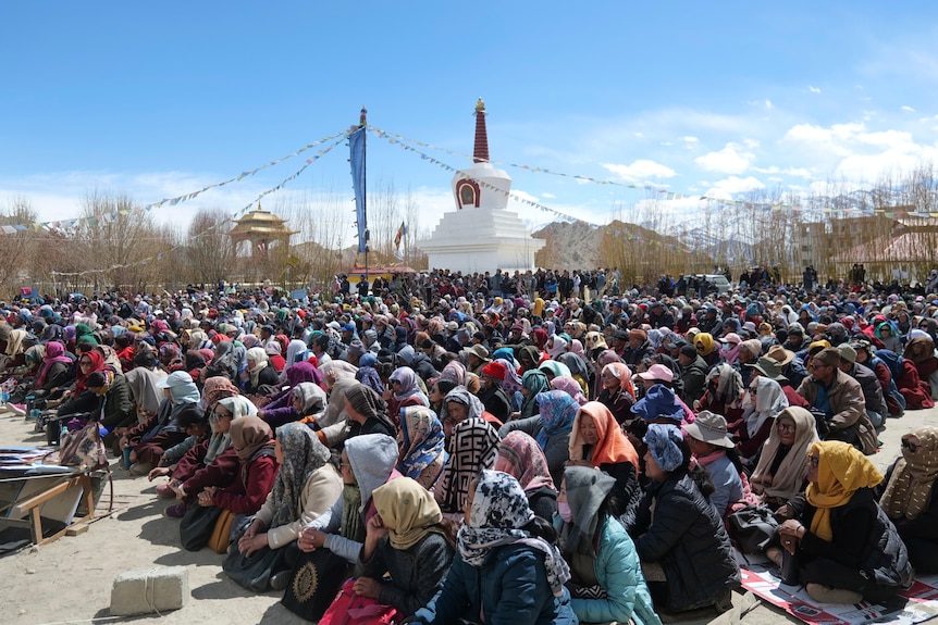 Thousands of people in shawls sit on the ground near a monument under blue sky