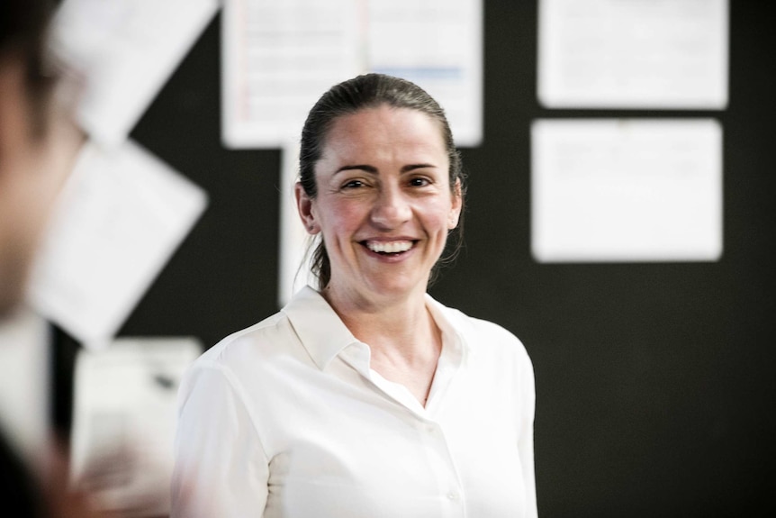 Lee Lewis wearing white shirt and with her dark brown hair in a ponytail smiling at camera, notice board in background.