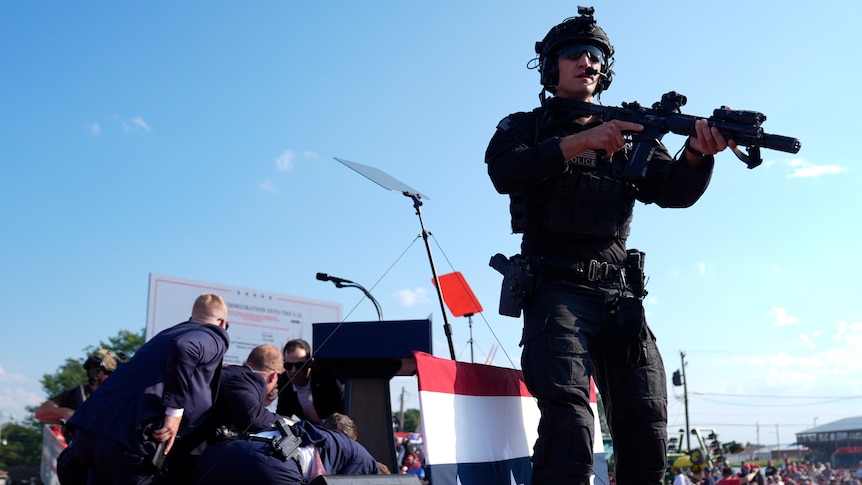 A soldier in black with an assault rifle looms over the stage with a pile of suited men covering Trump