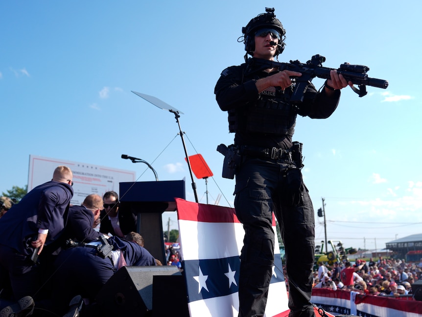 A soldier in black with an assault rifle looms over the stage with a pile of suited men covering Trump