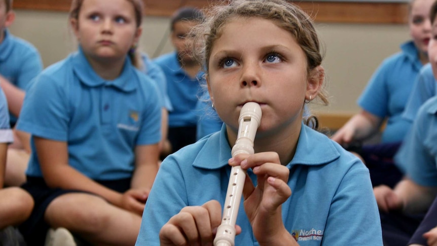 A young primary school aged girl plays a recorder in a classroom.