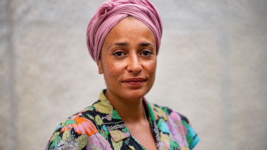 A woman with dark skin, a light pink turban and colourful patterned shirt looks at the camera