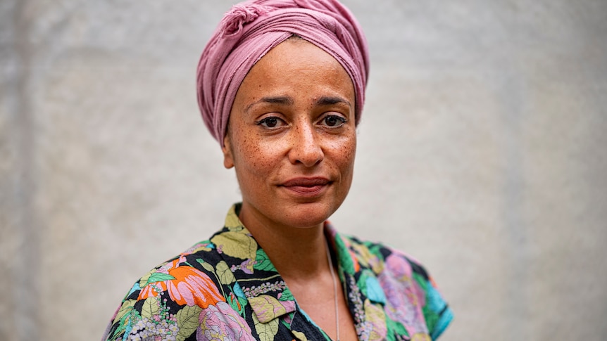 A woman with dark skin, a light pink turban and colourful patterned shirt looks at the camera