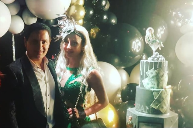 A man wearing a suit jacket and a woman in a sequined green dress pose next a large cake with balloons in the background