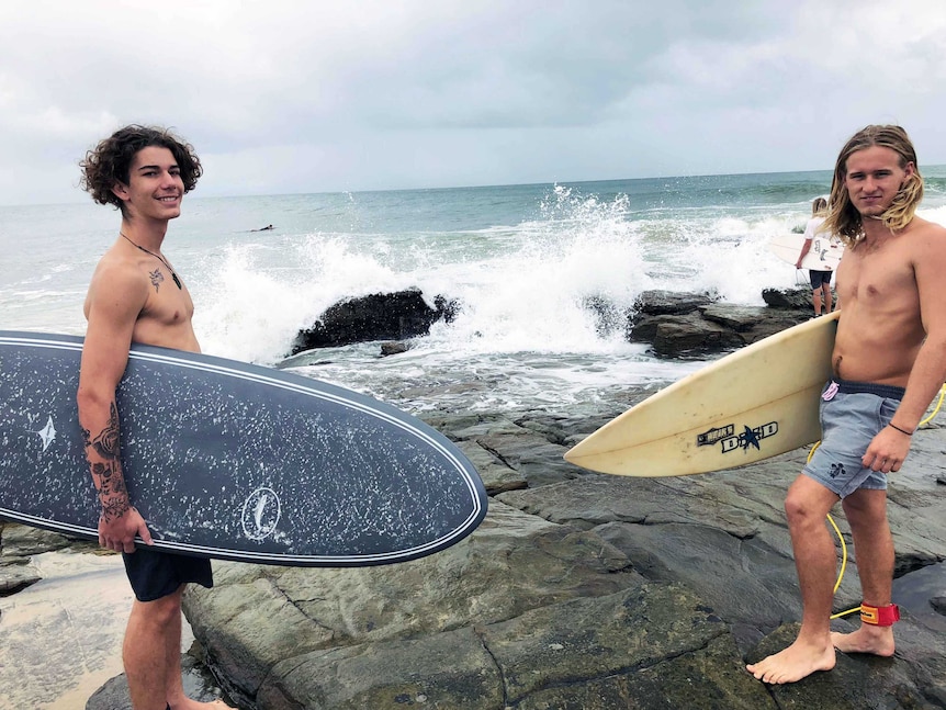 Mike McMaster and Max Wilson hold their surfboards in front waves on the rocks