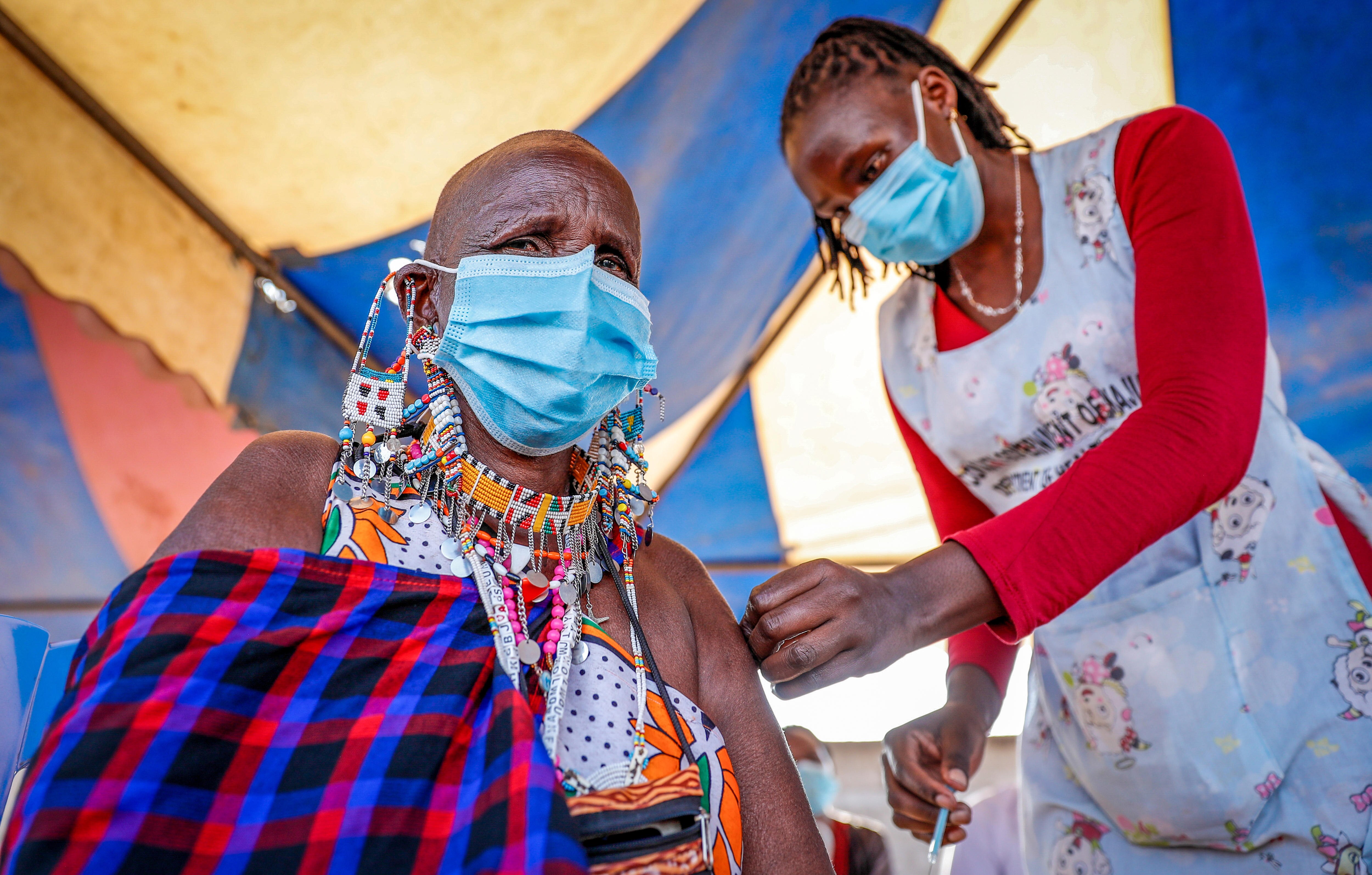 A Maasai woman in a blue face mask gets a needle injected in her arm by a female nurse