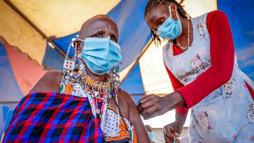 A Maasai woman in a blue face mask gets a needle injected in her arm by a female nurse