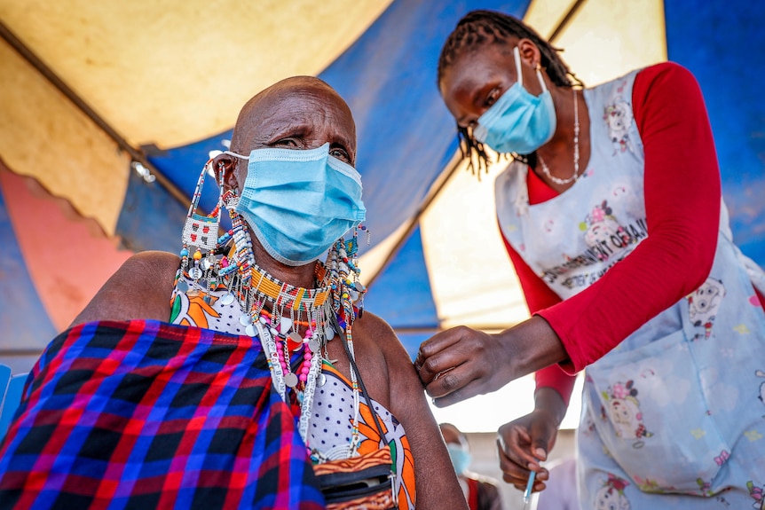 A Maasai woman in a blue face mask gets a needle injected in her arm by a female nurse