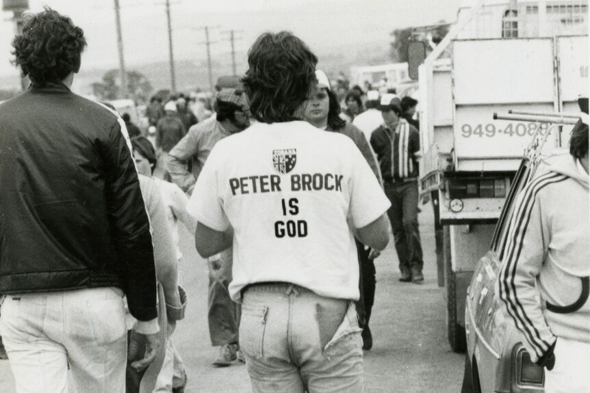 A man wearing a white t-shirt that says Peter Brock is God walks away from the camera.