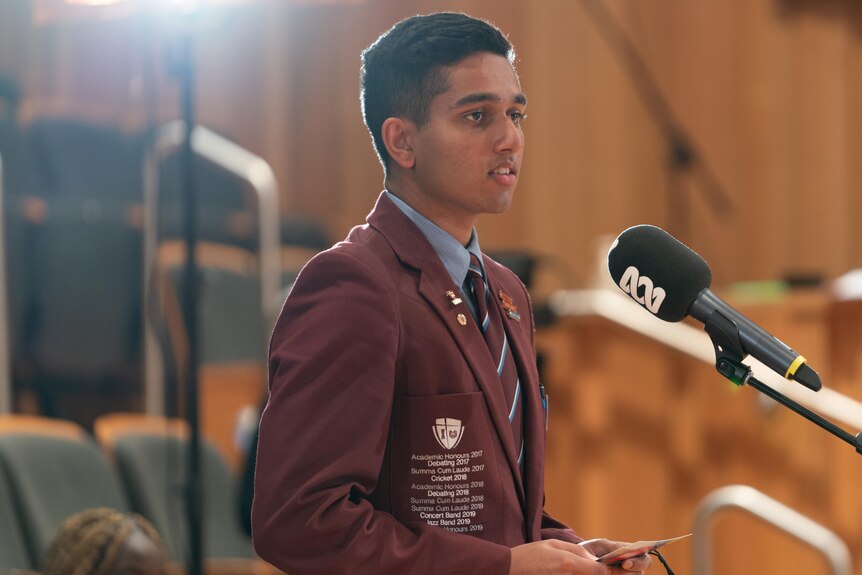 A male student with short black hair and a burgundy blazer stands at a microphone 