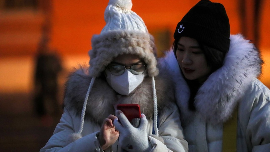 Two women, one wearing a mask, look at a smartphone.