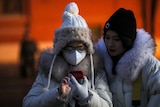 Two women, one wearing a mask, look at a smartphone.