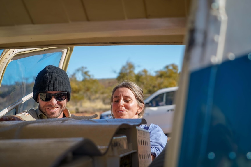 A man in a beanie and sunglasses and a woman in a blue shirt face boxes inside a helicopter.