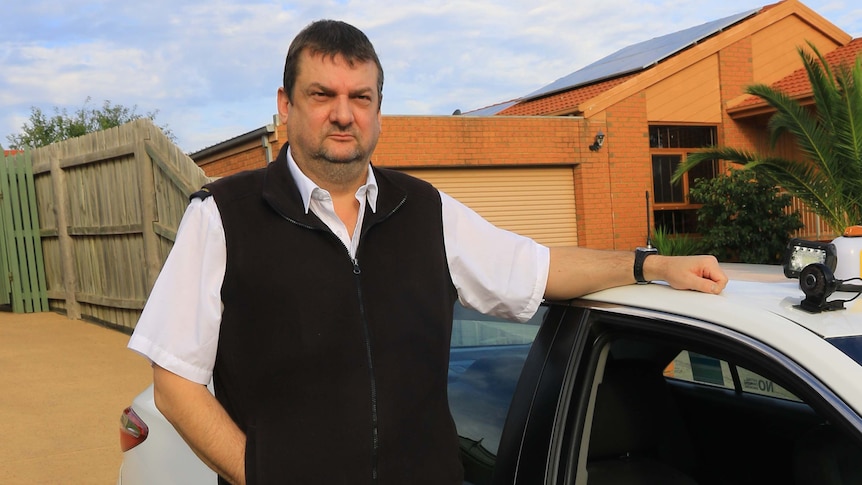 Paul Brooks, wearing a taxi uniform and standing next to his taxi with a serious expression on his face.
