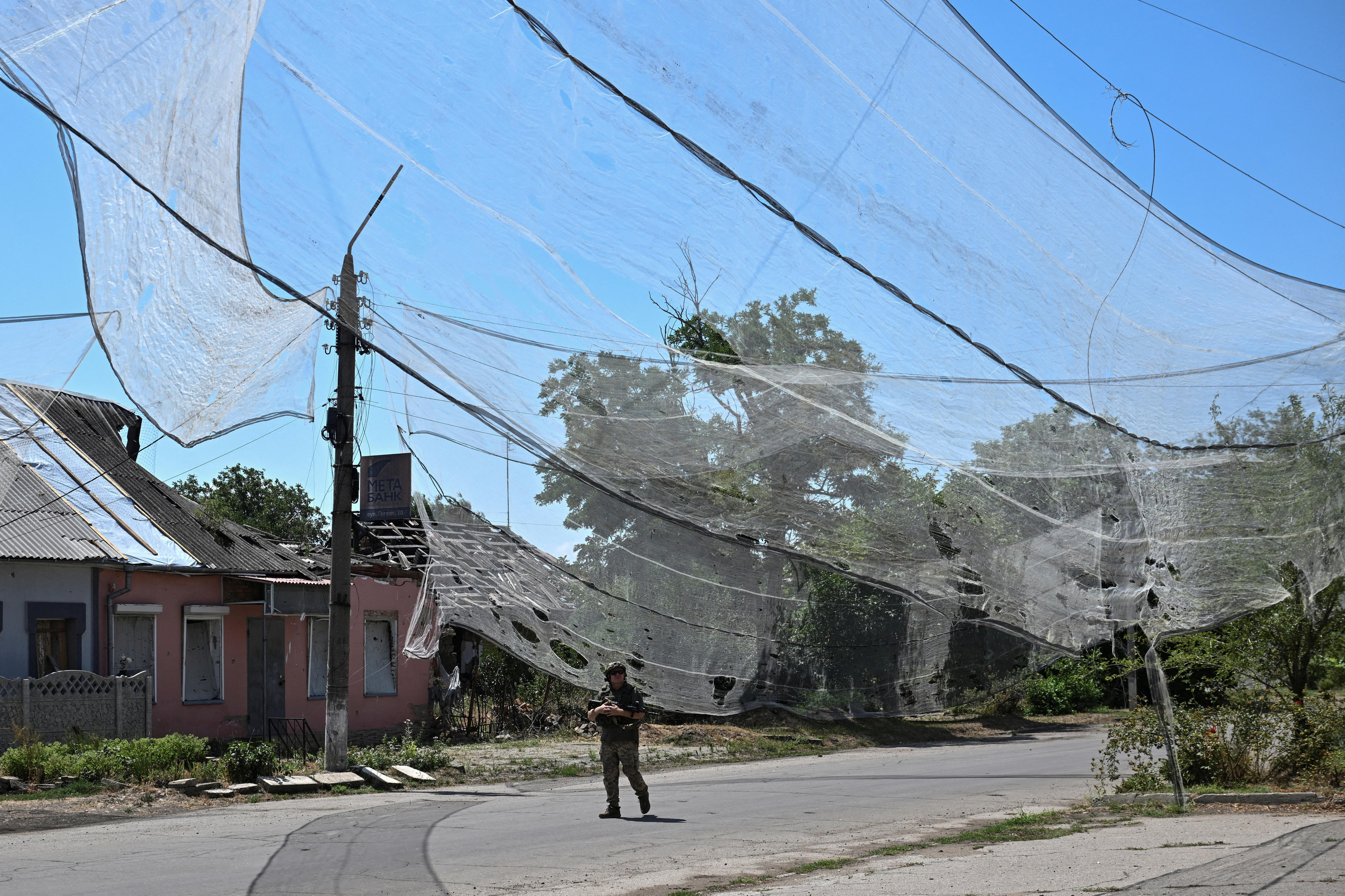 A soldier holds a gun and walks through a street where a huge, damaged net is draged across the whole street