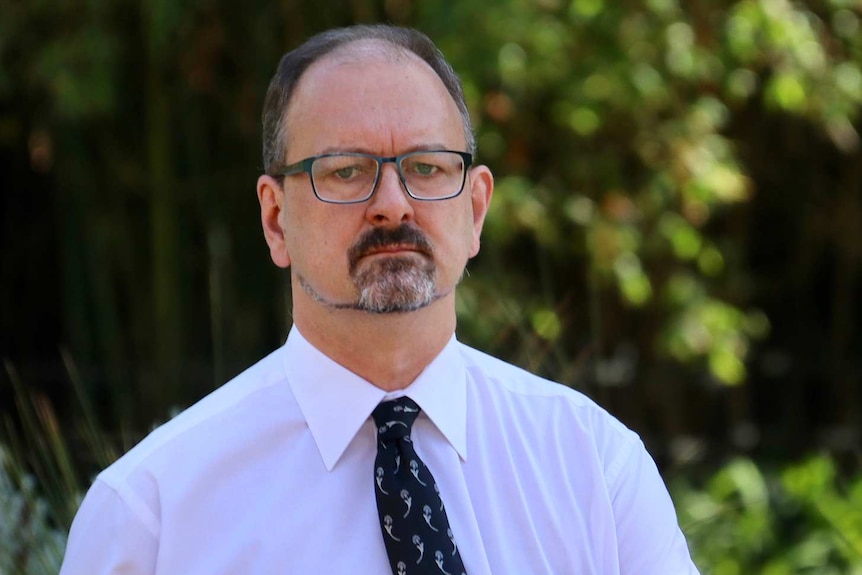 A man in a white shirt, black tie and glasses in front of some trees